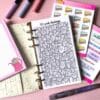 A personal ring planner open on a table. The top sheet shows a 'yearly saving challenge' insert. Around the planner are sticker sheets, a pink memo pad and some pens.