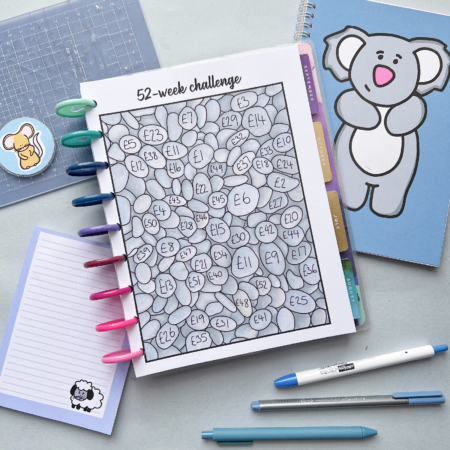 The 52 Week Saving Challenge 2 tracker features numbered heart shapes and is displayed with pens, a sheep notepad, a koala blue notebook, and a ruler on the desk.