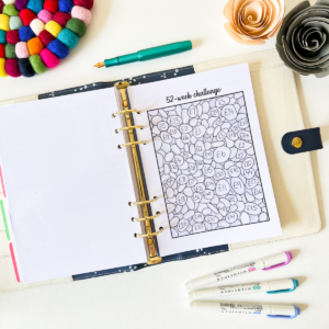 The 52 Week Saving Challenge planner is open to a numbered bubbles sheet, surrounded by colourful pens, felt circles, and paper flowers on a white desk.