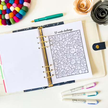 The 52 Week Saving Challenge planner is open to a numbered bubbles sheet, surrounded by colourful pens, felt circles, and paper flowers on a white desk.