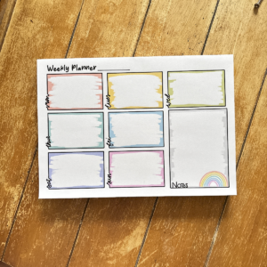 The A4 Full Weekly Planner Pad 3 features colourful boxes for each day, a spacious Notes section, and a rainbow illustration, all neatly arranged on a wooden surface.