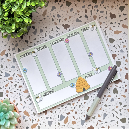 The A5 Bumblebee Weekly Planner Pad 3 features sections for weekdays, weekend, and notes with bee, flower, and beehive designs. A pen sits atop the pad beside two small potted plants on a speckled surface.