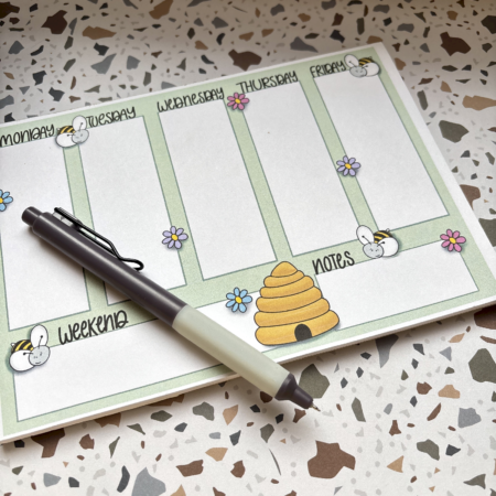 The A5 Bumblebee Weekly Planner Pad features bee and flower illustrations, sections for Monday to Friday, Weekend, and Notes, plus a beehive drawing. Shown with a black and white pen on a terrazzo background.