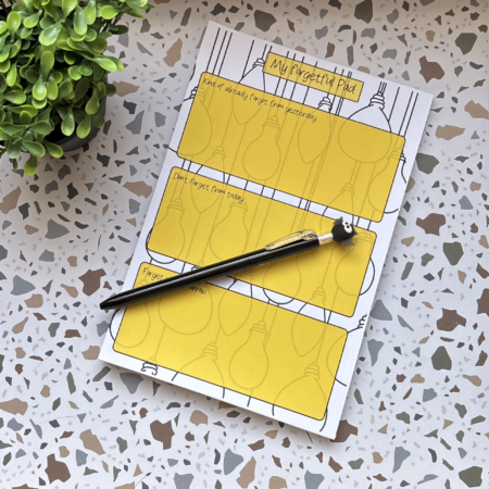 An A5 I Forget Things Memo Pad with yellow task sections, complemented by a black pen topped with a panda figure, sits beside a green plant on a terrazzo-patterned surface.