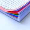 Close-up of the A5 Rainbow Priority Task List, showing a stack of colourful lined sheets—pink, blue, green, and white pages—slightly fanned out on a white surface.