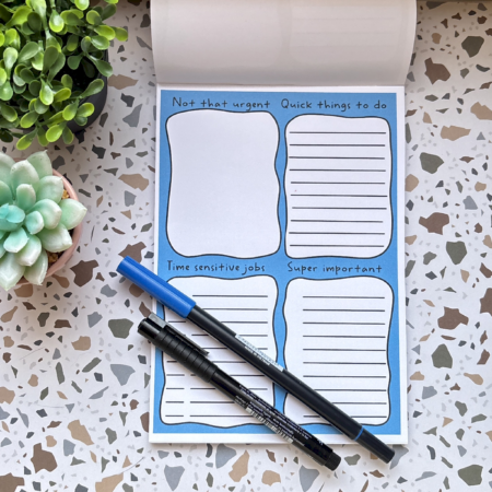 The A5 Rainbow Priority Task List 2, divided into four sections—Not that urgent, Quick things to do, Time sensitive jobs, and Super important—sits on a terrazzo surface with two pens and two small plants nearby.