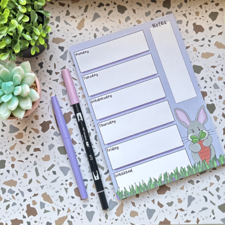 The A5 Weekly Planner Pad 2 features daily and notes sections, with a cute cartoon bunny holding a carrot. Shown near two pens and a succulent on a terrazzo-patterned surface.