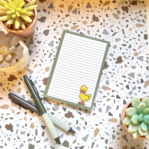 The A6 Worried Duck Memo Pad 2, featuring a yellow duck design, rests on a terrazzo-patterned surface with three potted succulents and three pens around it.
