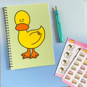 An A5 notebook with a spiral binding. The notebook has a pale green background and a duck illustration on it. Next to it is a teal fountain pen and two sticker sheets.