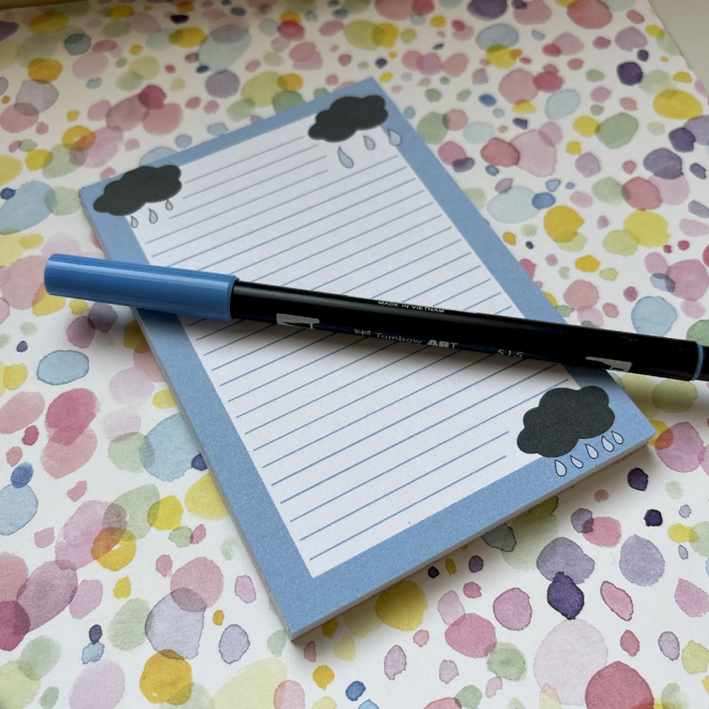 A blue pen rests on the April Showers A6 Memo Pad 2, featuring cloud designs in the corners, placed on a pastel spotty surface of pink, yellow, purple, and green.