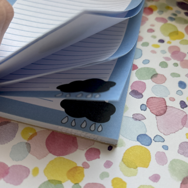 A close-up of a hand flicking through the April Showers A6 Memo Pad with blue-lined pages, each featuring a black rain cloud and raindrops—ideal for jotting notes—on a surface decorated with colourful paint splatters.