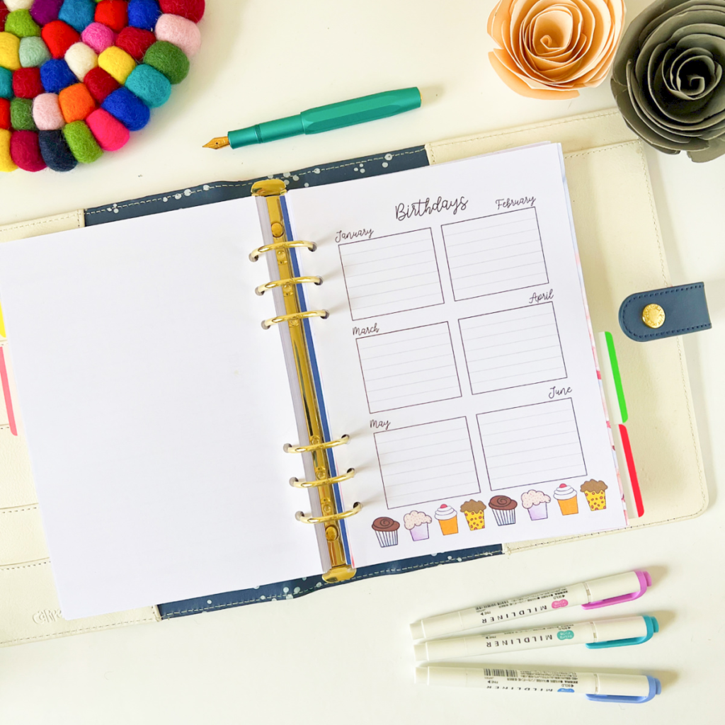 Birthday and Celebration Planner 3 is open to a birthday tracker for January–June, surrounded by colourful markers, a teal pen, and paper flowers on a white desk.