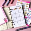 A personal ring planner open on a table. The top sheet shows a 'birthday year' insert. Around the planner are sticker sheets, a pink memo pad and some pens.