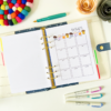 The Birthday Gift Planner Insert 2, decorated with cupcake illustrations, is displayed open on a white desk alongside colourful pens, a felt pom-pom coaster, and paper flowers.