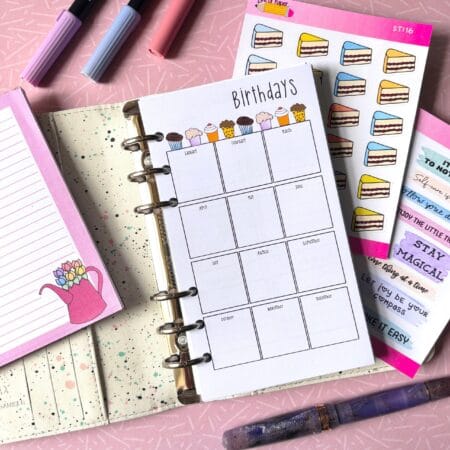 A personal ring planner open on a table. The top sheet shows a 'birthday year' insert. Around the planner are sticker sheets, a pink memo pad and some pens.