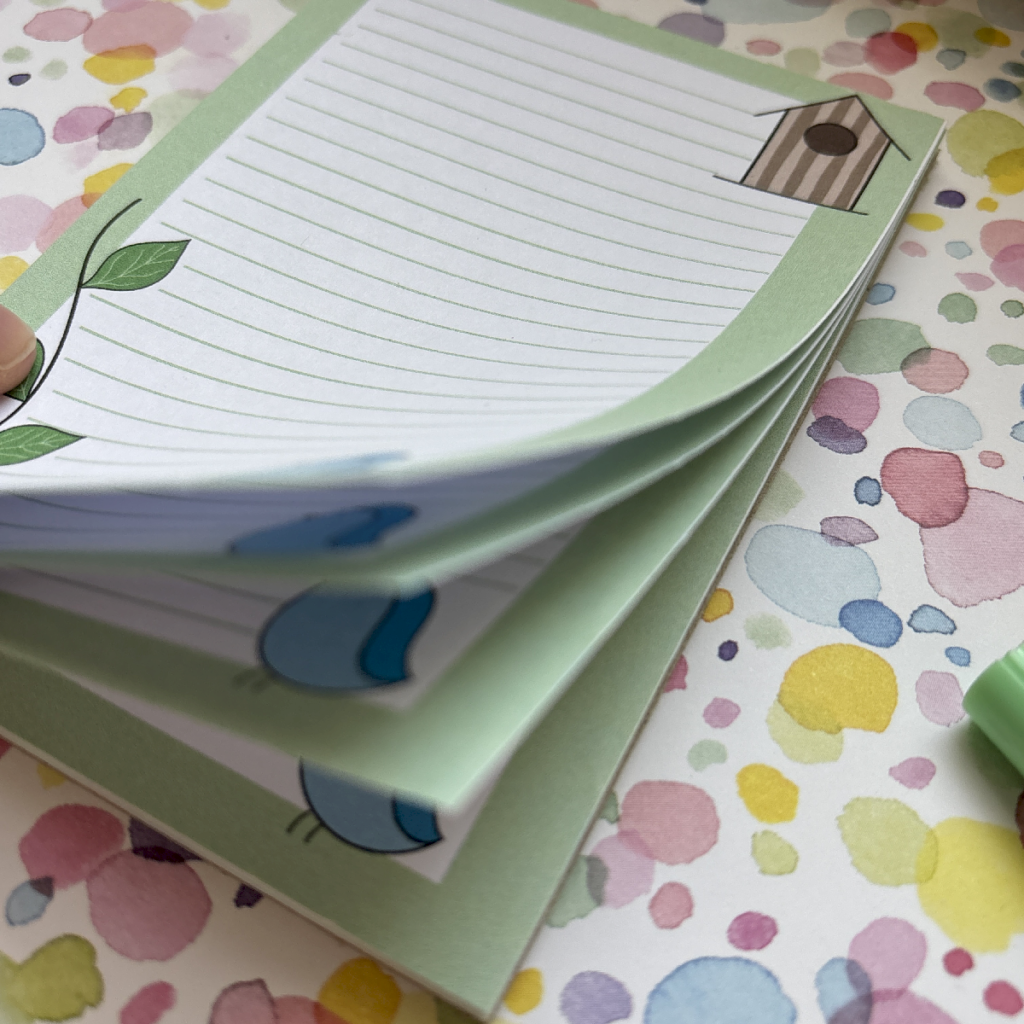 A hand flicks through green-lined pages from the Blue Bird A6 Memo Pad, featuring leaf and birdhouse designs, set against a colourful polka-dot background.