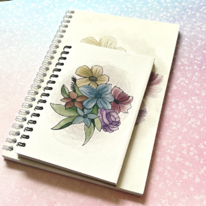 Two spiral bound notebooks lay on a pastel background. The notebooks have the same design, and the one on top is smaller than the bottom one. The notebook has a white background and an illustration of a bouquet of flowers in soft colours on it.