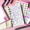 A personal ring planner open on a table. The top sheet shows a 'colouring sheet' insert. Around the planner are sticker sheets, a pink memo pad and some pens.