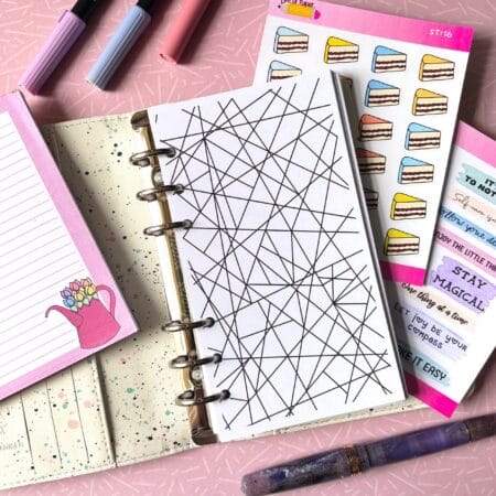A personal ring planner open on a table. The top sheet shows a 'colouring sheet' insert. Around the planner are sticker sheets, a pink memo pad and some pens.