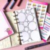 A personal ring planner open on a table. The top sheet shows a 'colouring sheet' insert. Around the planner are sticker sheets, a pink memo pad and some pens.