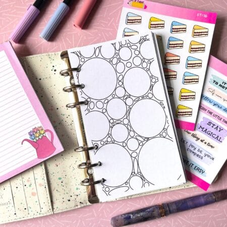 A personal ring planner open on a table. The top sheet shows a 'colouring sheet' insert. Around the planner are sticker sheets, a pink memo pad and some pens.