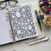 An open Colouring Sheet Planner Insert 6 displays a page with overlapping black circles. Surrounding it are planner sheets, glasses, pens, a felt pom-pom coaster, a paper flower, and white markers on a white desk.