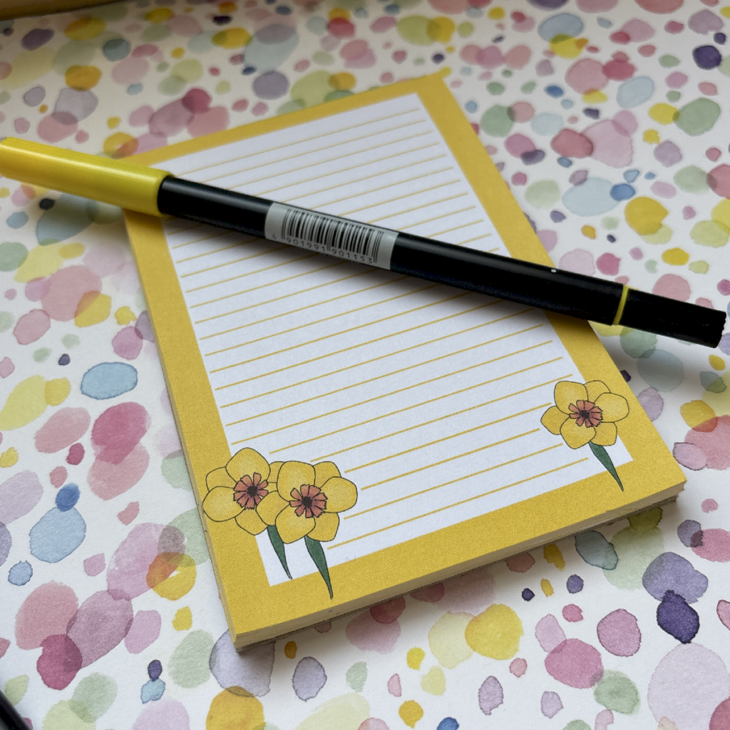 A black and yellow pen rests on the Daffodil A6 Memo Pad 2, decorated with yellow flower illustrations, and placed on a colourful, spotted surface.