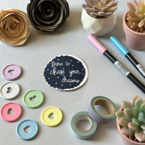 A light grey background with a single sticker lay in the centre. A large deep blue circlular sticker. In white there are little stars dotted across it and the white text says 'dare to chase your dreams'. Around the edge are paper roses, washi tape, planner discs and brush pens.