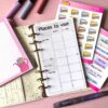 A personal ring planner open on a table. The top sheet shows a 'places to visit' insert. Around the planner are sticker sheets, a pink memo pad and some pens.