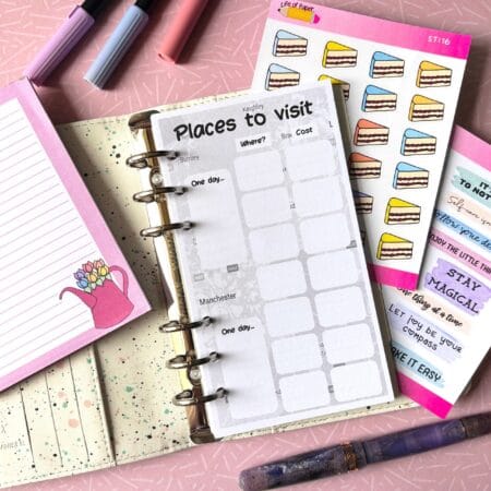 A personal ring planner open on a table. The top sheet shows a 'places to visit' insert. Around the planner are sticker sheets, a pink memo pad and some pens.