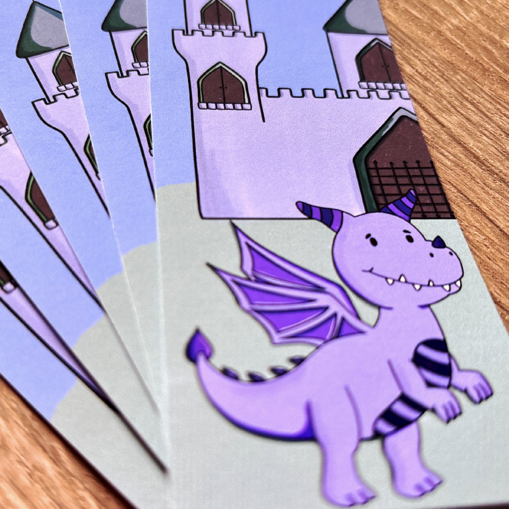 A close-up of Img 0583, children's adventure bookmarks featuring a smiling purple dragon with wings and striped horns standing before a castle, all set on a wooden surface.