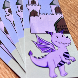 A close-up of Img 0583, children's adventure bookmarks featuring a smiling purple dragon with wings and striped horns standing before a castle, all set on a wooden surface.
