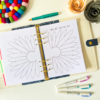 The Gratitude Flower Planner Insert 2 is open on a white desk, featuring a “What am I grateful for?” prompt and two large daisy outlines, surrounded by colourful pens, felt pom-poms, and paper flowers.