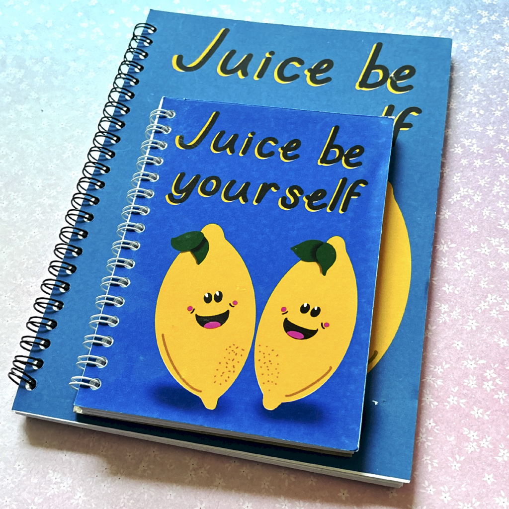 Two spiral bound notebooks lay on a pastel background. The notebooks have the same design, and the one on top is smaller than the bottom one. The notebook has a dark blue background and an illustration of two lemons with happy faces on them. Above them is the phrase 'juice be yourself'.