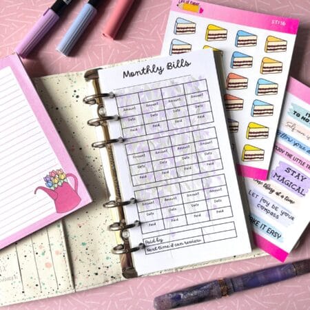 A personal ring planner open on a table. The top sheet shows a 'monthly bills' insert. Around the planner are sticker sheets, a pink memo pad and some pens.