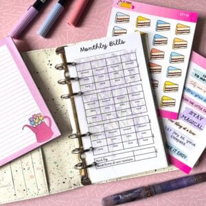 Monthly Bills Personal Ring Insert 2 A personal ring planner open on a table. The top sheet shows a 'monthly bills' insert. Around the planner are sticker sheets, a pink memo pad and some pens.