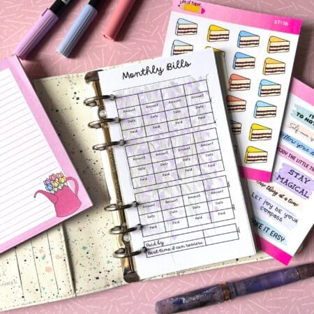 A personal ring planner open on a table. The top sheet shows a 'monthly bills' insert. Around the planner are sticker sheets, a pink memo pad and some pens.