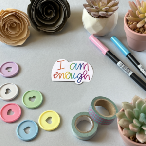 A light grey background with a single sticker lay in the centre. A sticker with a white background and just text that reads 'I am enough'. The writing fades through the colours if the rainbow from left to right. Around the edge are paper roses, washi tape, planner discs and brush pens.