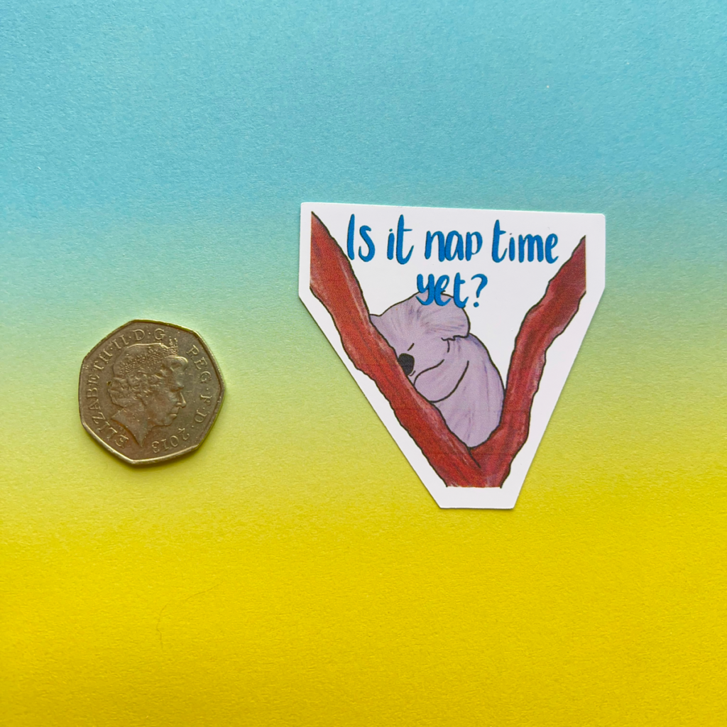 A 50 pence coin is placed beside the Is It Nap Time Matt Vinyl Sticker, featuring a smiling sloth on a tree branch with “Is it nap time yet?” above it on a blue and yellow gradient background.