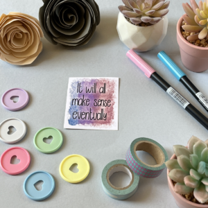 A light grey background with a single sticker lay in the centre. A rectangular sticker with a muted purple and grey marbled background The text in black says 'it will all make sense eventually'. Around the edge are paper roses, washi tape, planner discs and brush pens.