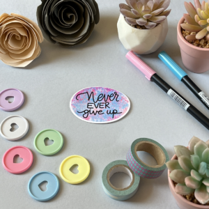 A light grey background with a single sticker lay in the centre. An oval shaped sticker with a marbled pink and blue design and the text in black reads 'never ever give up'. Around the edge are paper roses, washi tape, planner discs and brush pens.