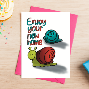 New Home Greeting Card 4 comes in a pink envelope and features two illustrated snails with the message “Enjoy your new home.” The card is pictured on a wooden table beside a cupcake, hundreds and thousands, and a blue gift box.