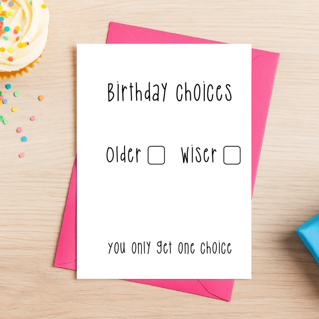 A pink envelope holds the Older Or Wiser Birthday Card 4, which reads: “Birthday Choices. Older ☐ Wiser ☐ You only get one choice.” It rests on a wooden table with a cupcake and confetti nearby.