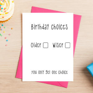A pink envelope holds the Older Or Wiser Birthday Card 4, which reads: “Birthday Choices. Older ☐ Wiser ☐ You only get one choice.” It rests on a wooden table with a cupcake and confetti nearby.