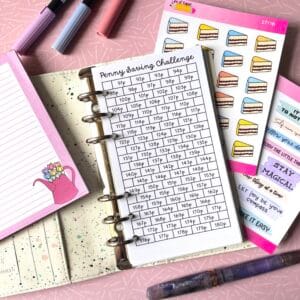 A personal ring planner open on a table. The top sheet shows a 'penny challenge' insert. Around the planner are sticker sheets, a pink memo pad and some pens.