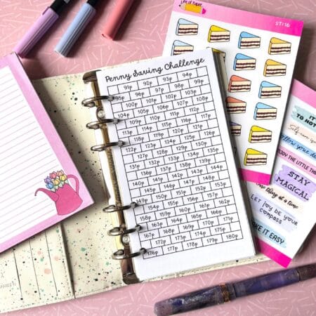 A personal ring planner open on a table. The top sheet shows a 'penny challenge' insert. Around the planner are sticker sheets, a pink memo pad and some pens.