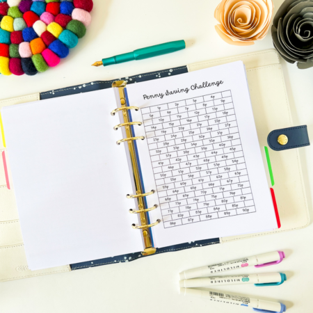 An A5 6-ring planner is open on a table. The top sheet shows the penny savings challenge. This has brick wall outlines with the bricks numbered from 1p to 365p. The top of each sheet says 'penny savings challenge'. The picture has a cream background. Around the planner is a rainbow pompom coaster, roses made from card and highlighter pens. 