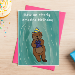The Otter Birthday Card 4 features a cartoon otter in a party hat holding a present and floating on its back. The card says "Have an otterly amazing birthday" and is shown with a pink envelope beside a fairy cake and confetti.