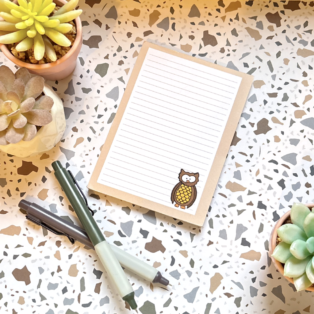 The Owl A6 Memo Pad 2, with a cute owl illustration in the bottom corner, is placed on a terrazzo-patterned surface alongside three potted succulents and green and grey pens.