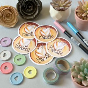 A light grey background with a pile of stickers in the centre. All the stickers have the same design. A circular sticker with a birds eye view of a latte and swirls of the coffee to make latte art. The text reads 'pause, breathe'. Around the edge are paper roses, washi tape, planner discs and brush pens.